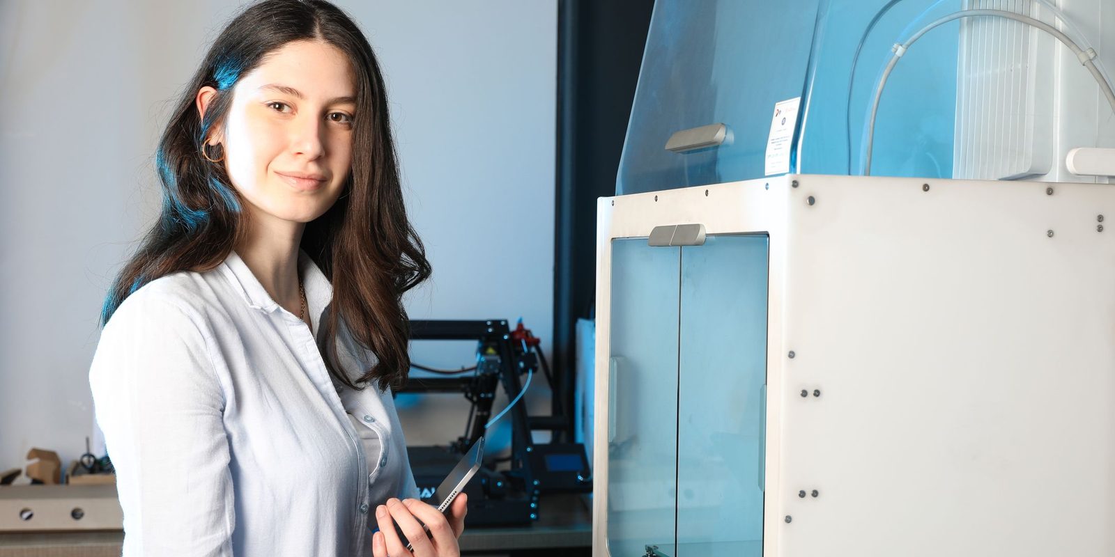 An engineer in a white shirt holds a tablet beside an enclosed 3D printer