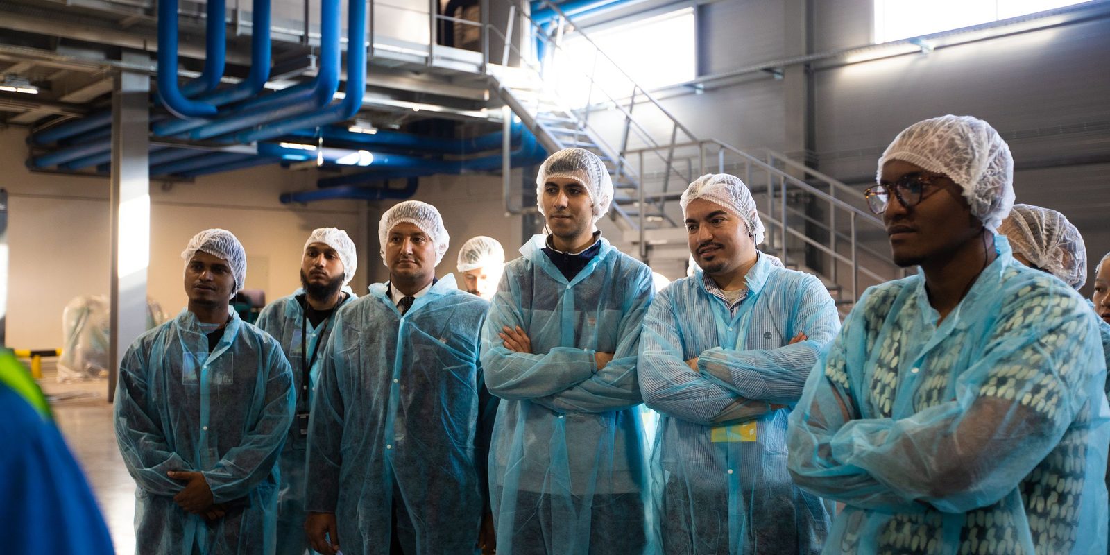 A group of visitors in disposable coats and hairnets stands on the floor of a bright plant with blue pipework overhead