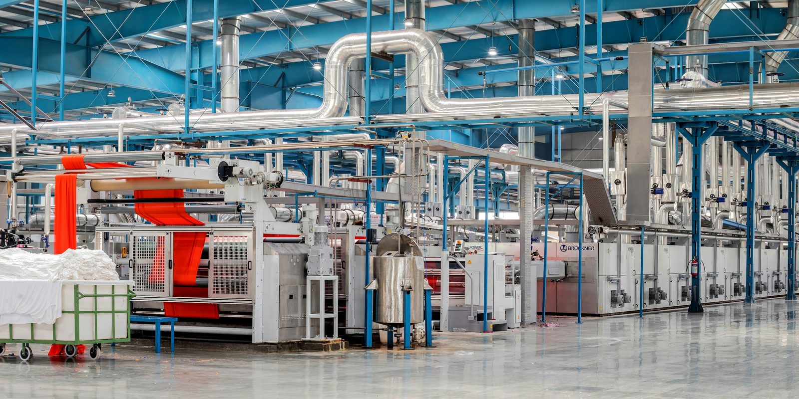 Fabric runs through finishing machines in a bright textile plant, under silver ducts and blue steel framing