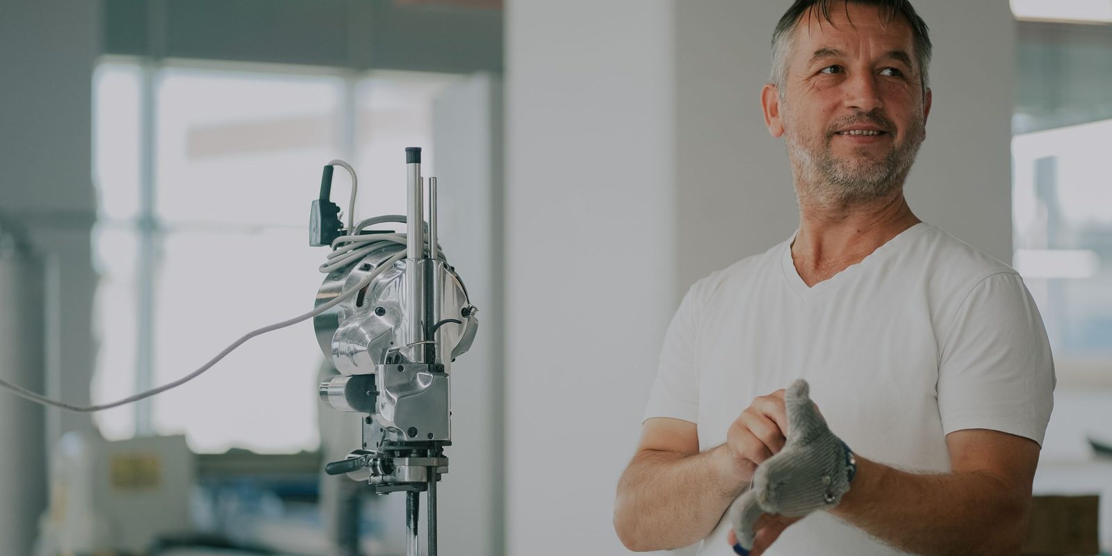 A man in a white T-shirt pulls on a work glove beside a fabric cutting machine in a bright workshop
