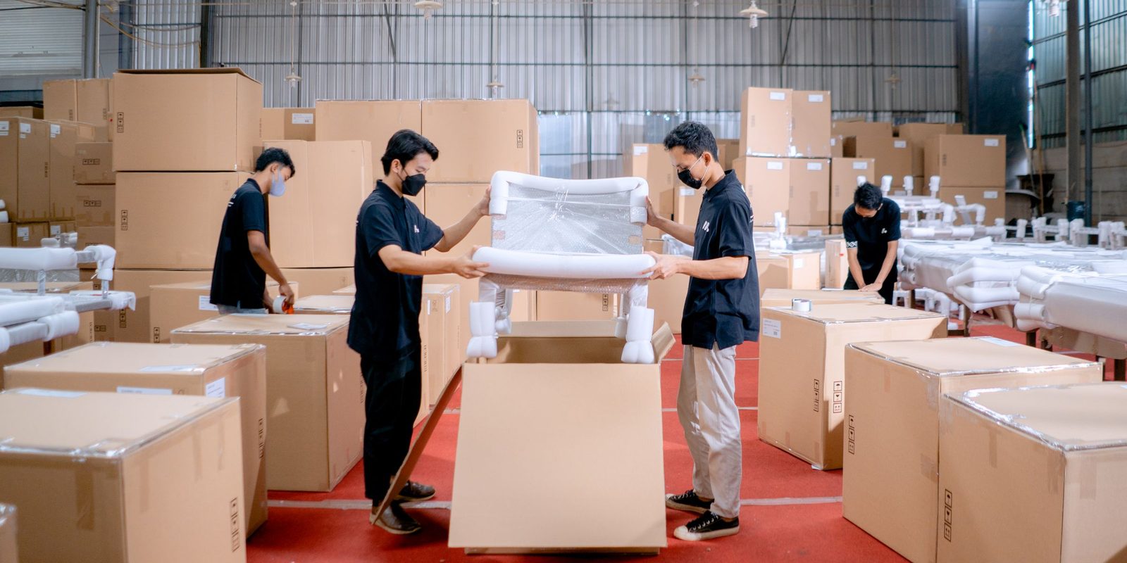 Workers in face masks lower a chair wrapped in foam into a shipping box, among stacks of boxes in a warehouse