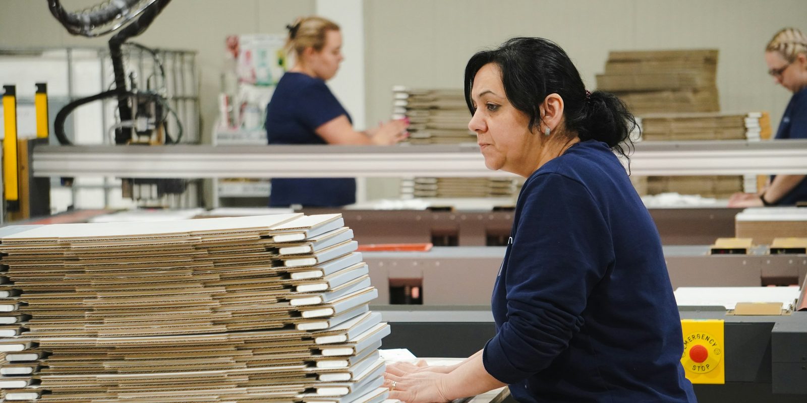 A worker squares a stack of flat cardboard boxes at a box-making machine in a bright packaging plant, with two colleagues behind her