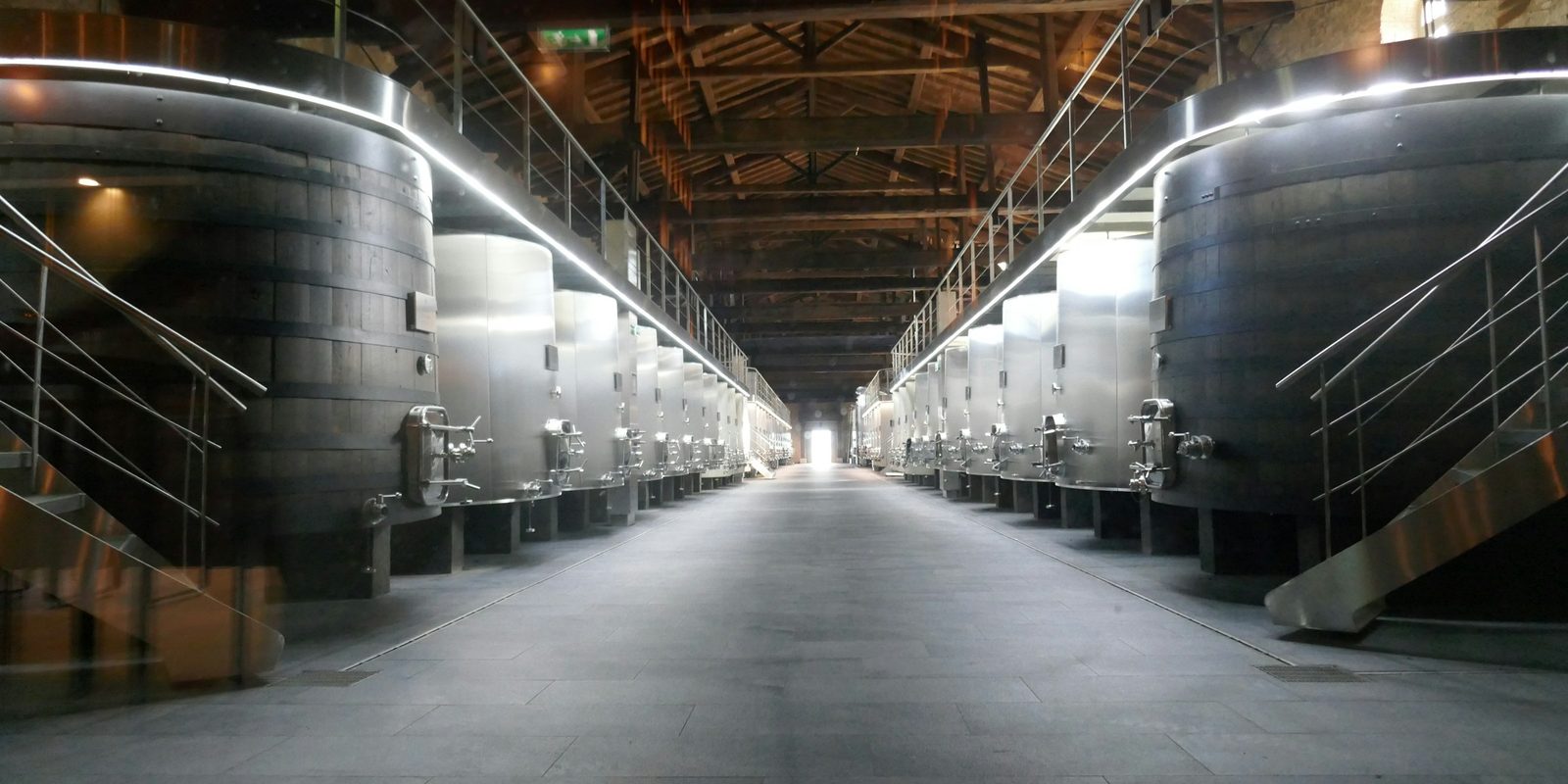 Large steel storage tanks line both sides of a long, lit aisle in a wine cellar
