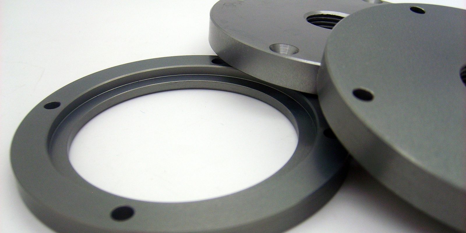 Two machined grey metal flanges and a ring with bolt holes lie on a white surface