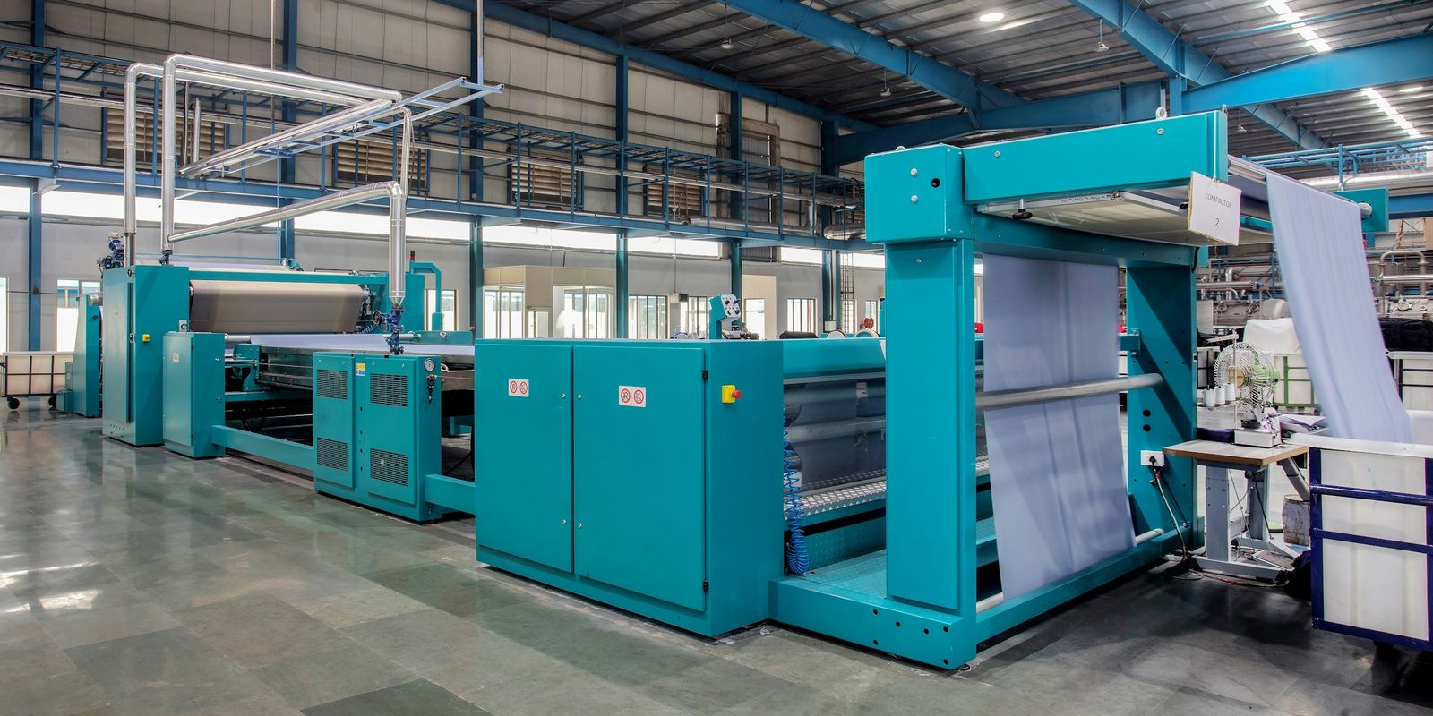 A long teal fabric compactor feeds pale cloth through rollers on a polished floor in a bright textile plant