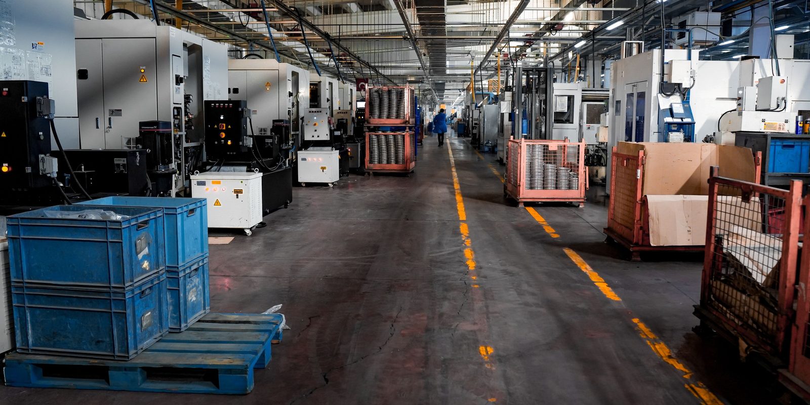 A long aisle on a machining floor lined with CNC machines, with blue parts bins on a pallet and wire baskets of finished parts