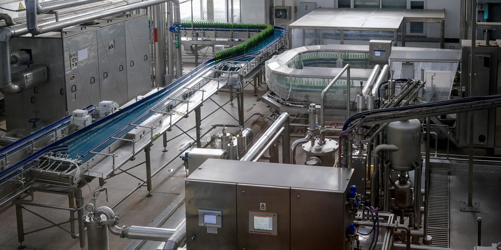 A bright bottling hall with conveyors, stainless steel control cabinets with touchscreens, and green bottles on the line