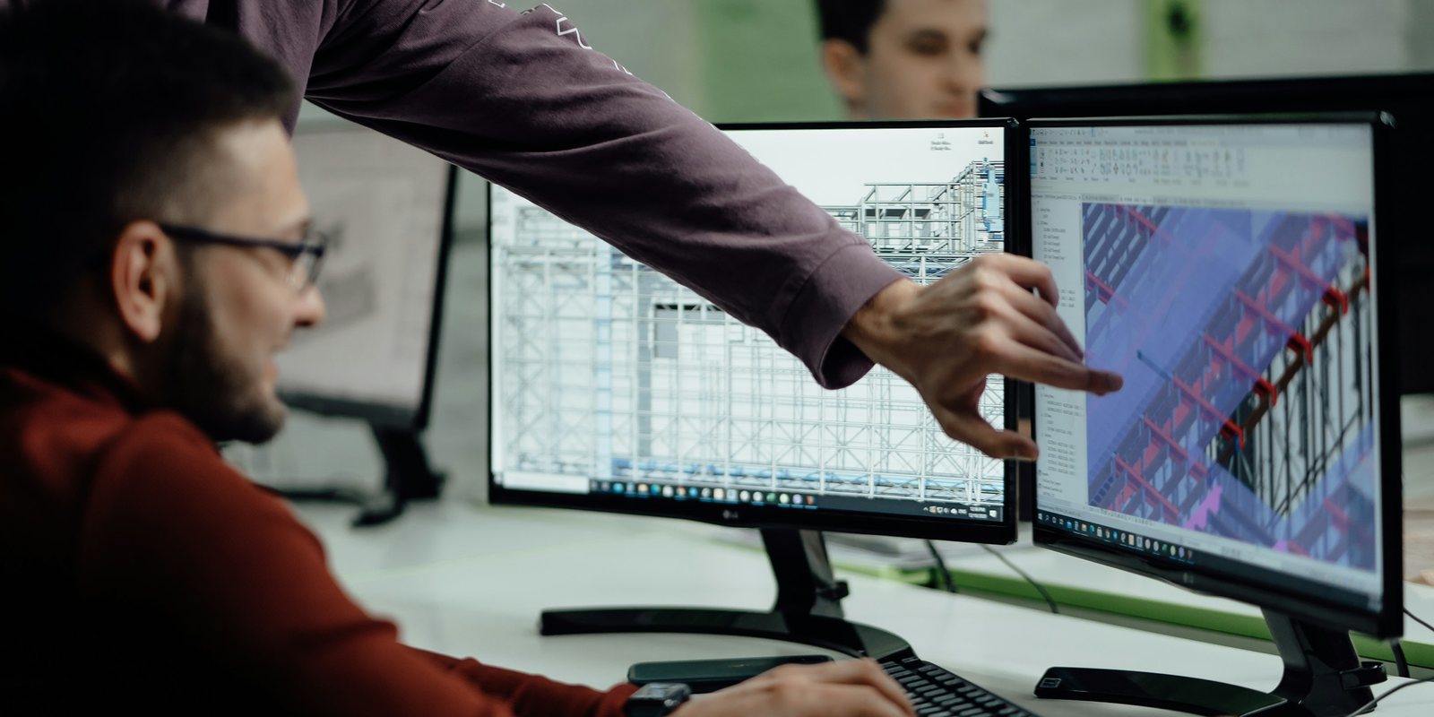 Two engineers at a desk review a 3D steel structure model on two monitors, one pointing at the screen