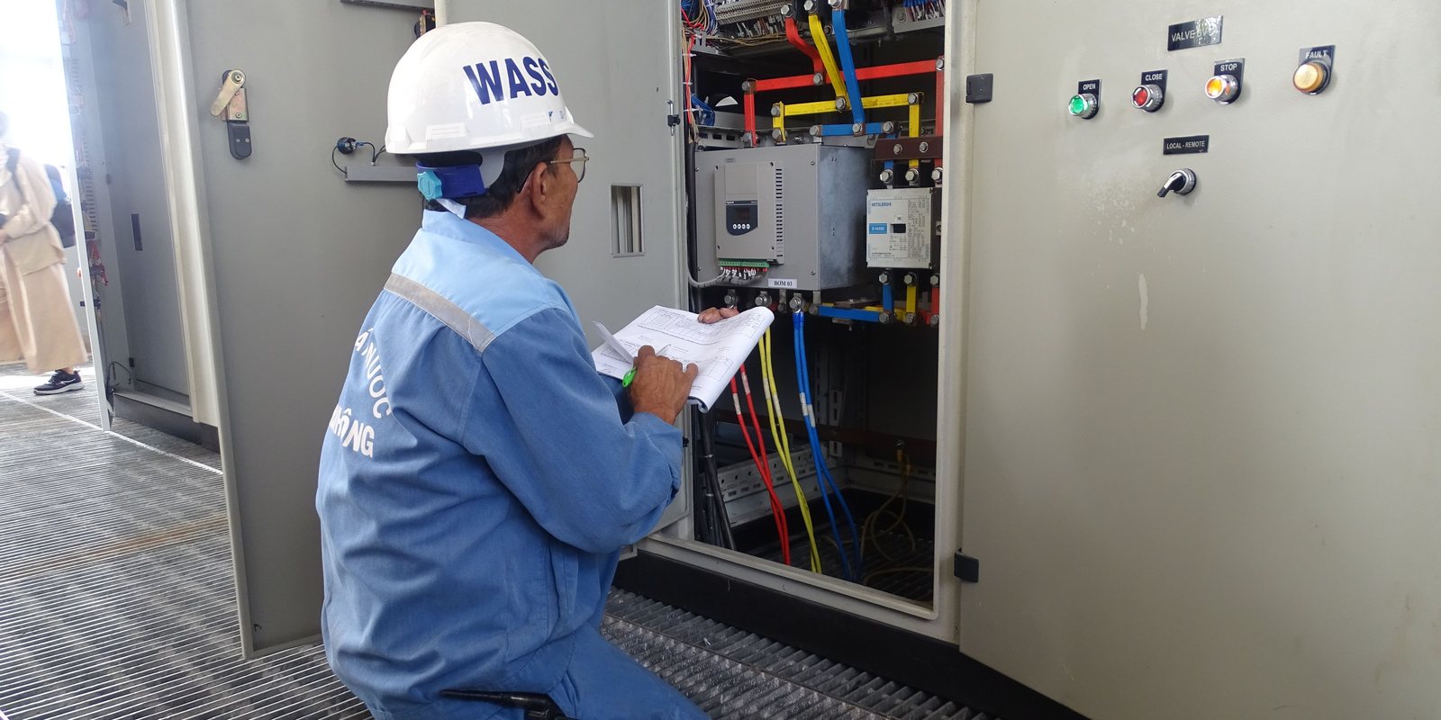 An engineer in a white hard hat kneels at an open electrical control cabinet, checking the wiring against a printed checklist