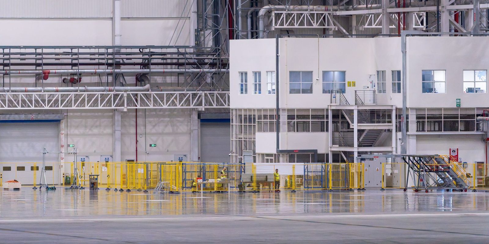 A clean, brightly lit production hall with a polished floor, yellow safety fencing and a two-storey office block