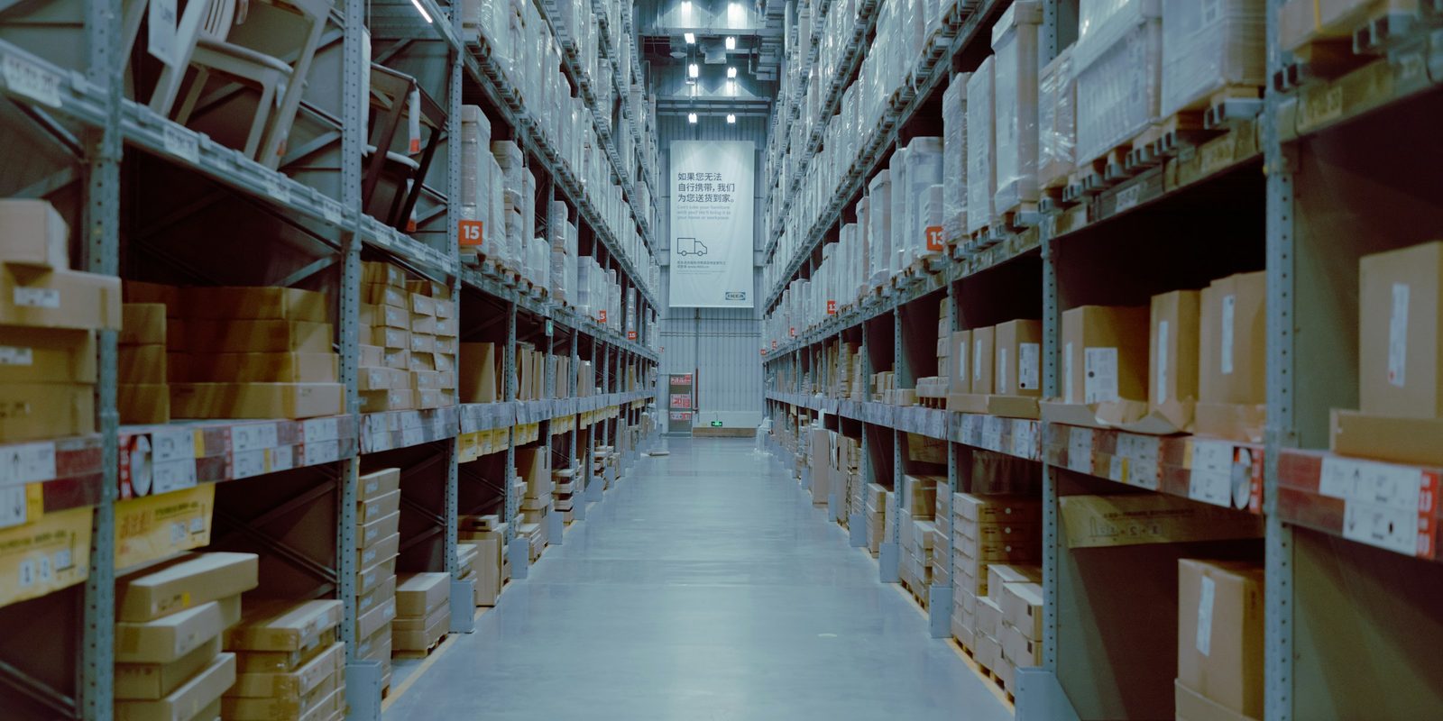 A long, bright warehouse aisle with tall shelves of boxed stock on both sides
