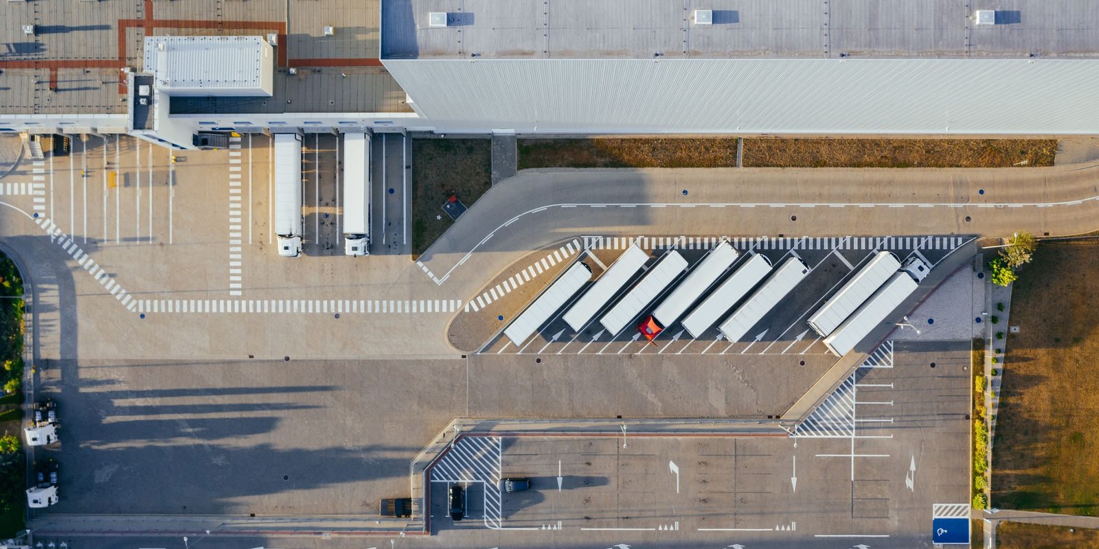 Aerial view of a modern logistics centre with white trailers parked in a row beside the loading docks and a large grey warehouse roof