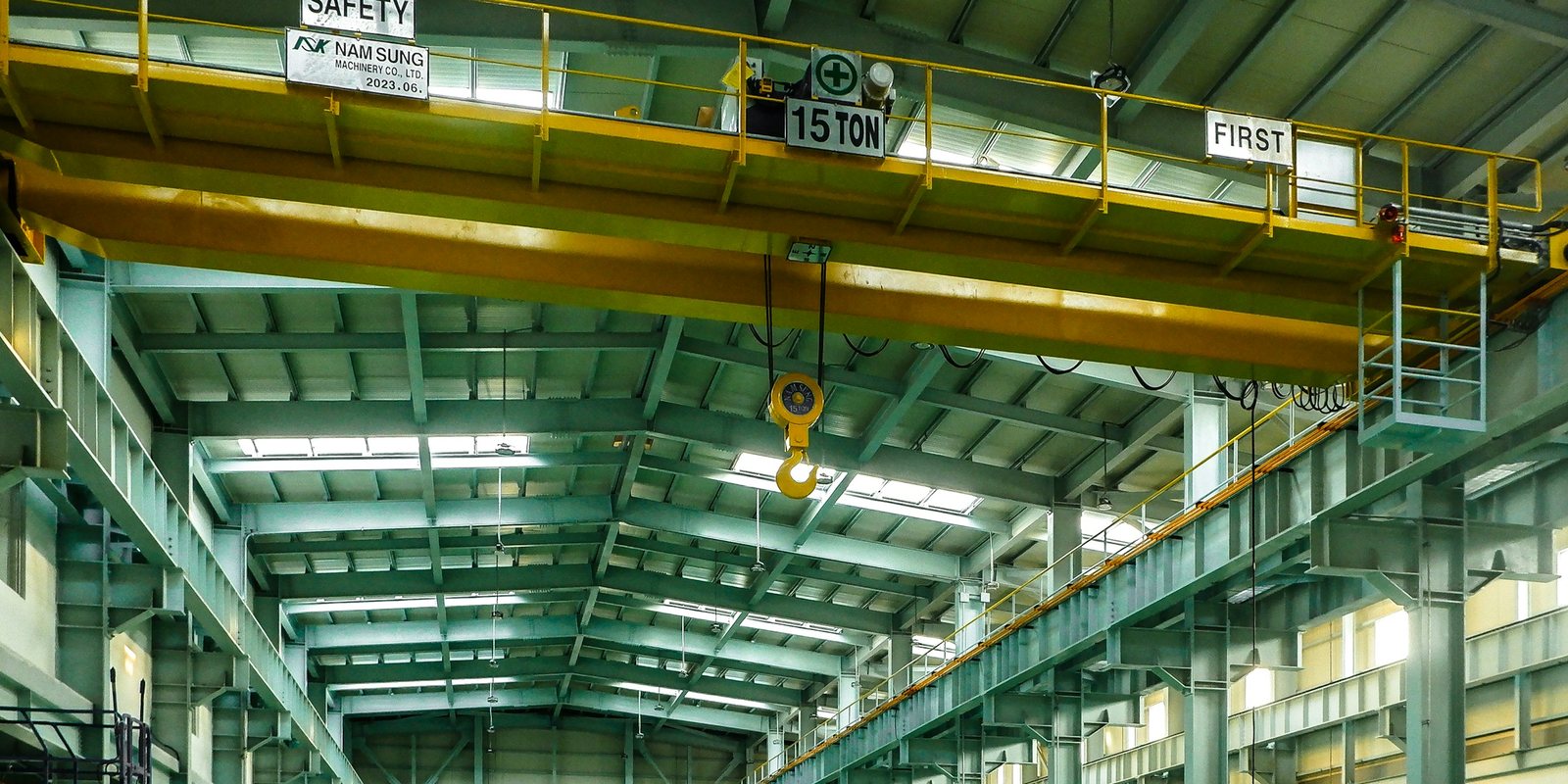 An empty, brightly lit assembly hall with green steel columns and a yellow 15-tonne overhead crane