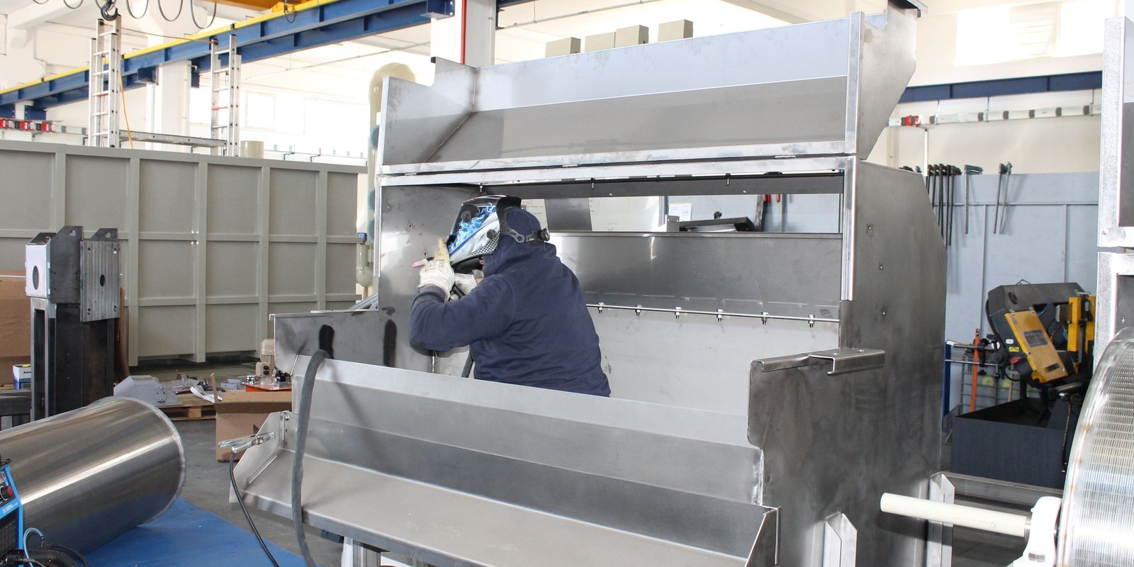 A welder in a protective mask builds a custom stainless steel machine frame in a bright fabrication shop with an overhead crane
