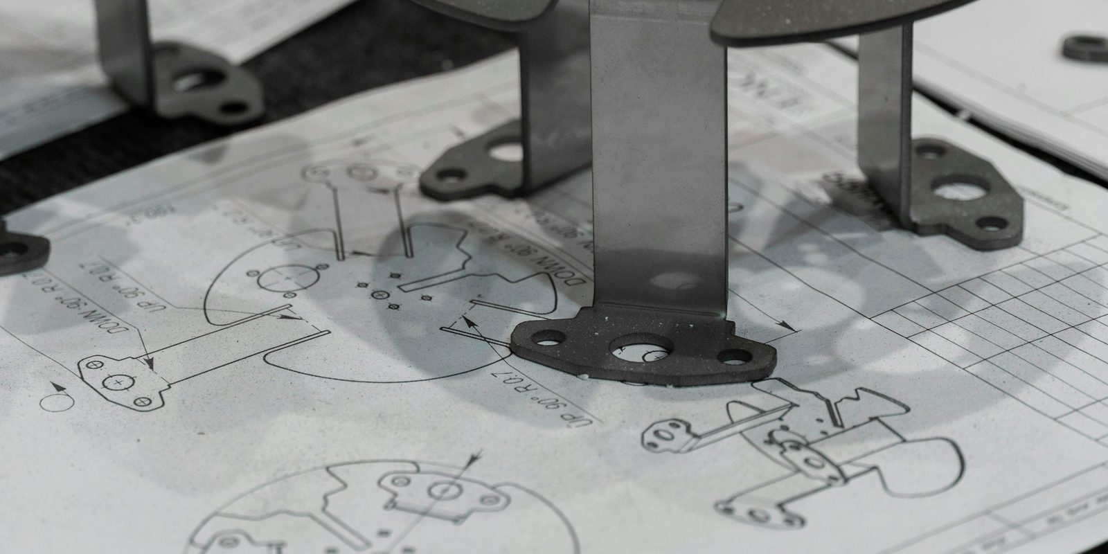 A bent sheet metal bracket resting on printed engineering drawings with dimensions and hole callouts