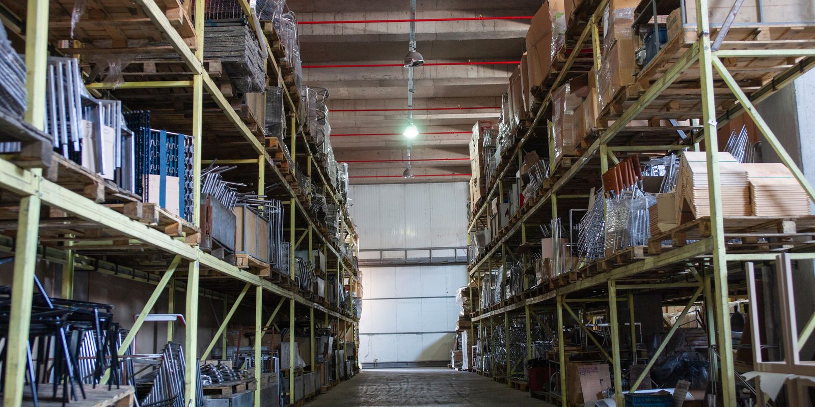 A long, well-lit warehouse aisle between tall green pallet racks stocked with boxed and palletized building materials