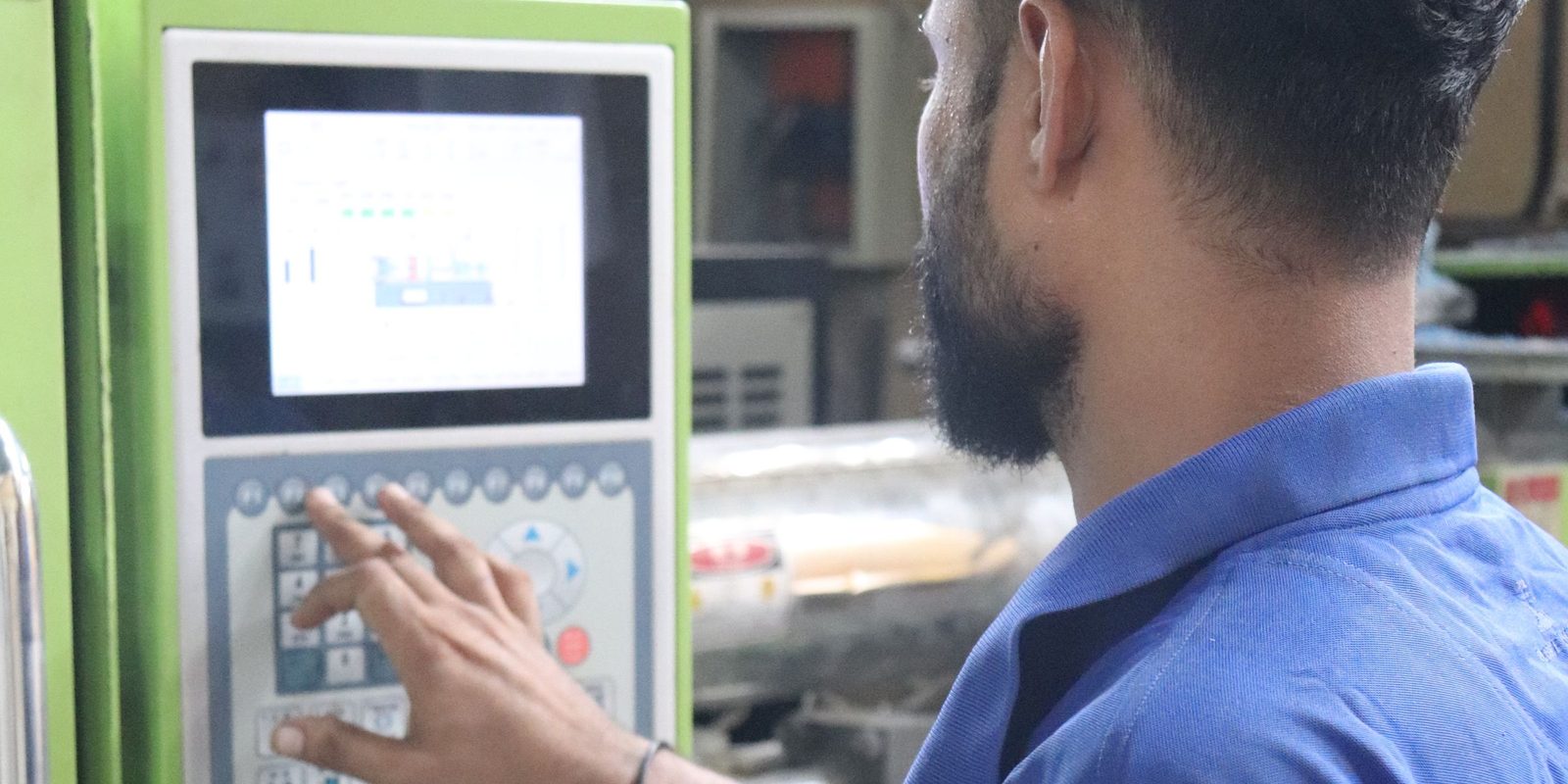 Un travailleur en chemise bleue appuie sur les touches du panneau de commande d'une machine de moulage verte, sous un &eacute;cran d'&eacute;tat