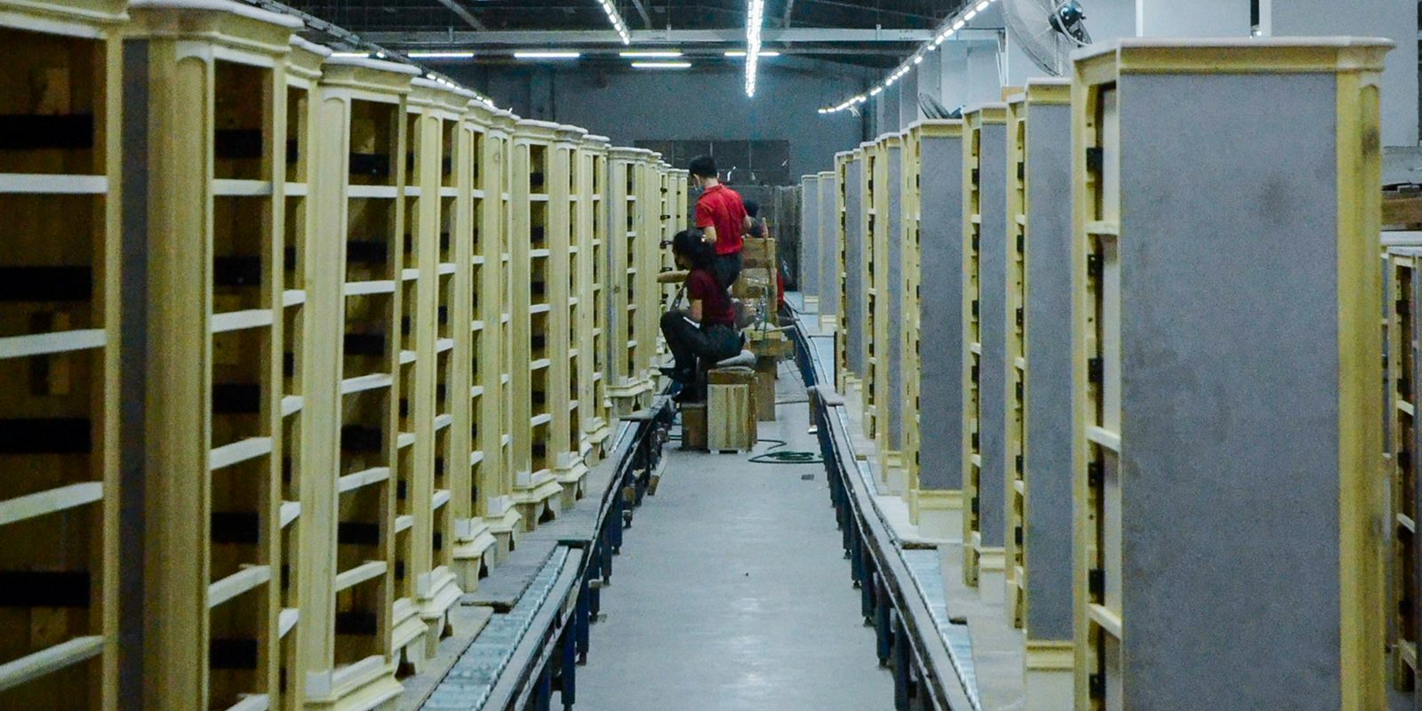 Une longue ligne de montage d'armoires en bois sur des convoyeurs &agrave; rouleaux, avec deux travailleurs au fond, dans une usine de meubles