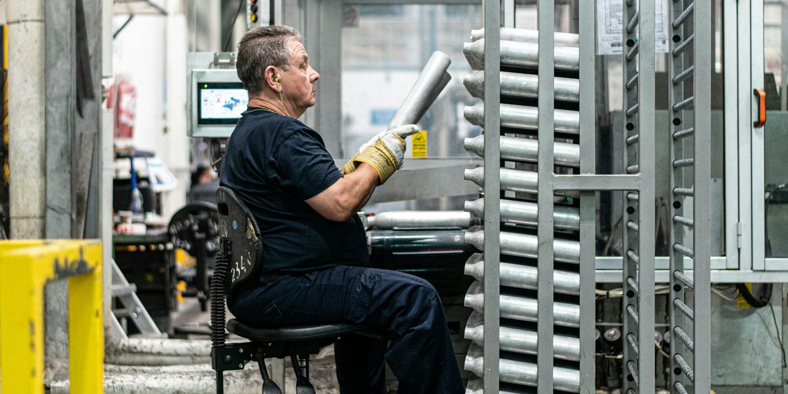 Un travailleur assis charge des tubes d'aluminium dans un support roulant, pr&egrave;s d'une machine automatis&eacute;e munie d'un &eacute;cran tactile, dans une usine &eacute;clair&eacute;e