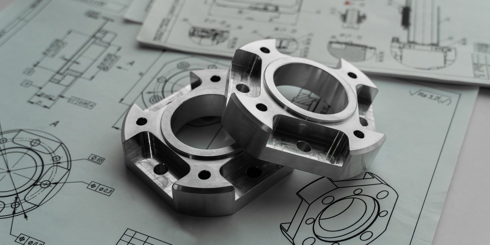 Two machined aluminium brackets resting on printed engineering drawings with section views and dimensions