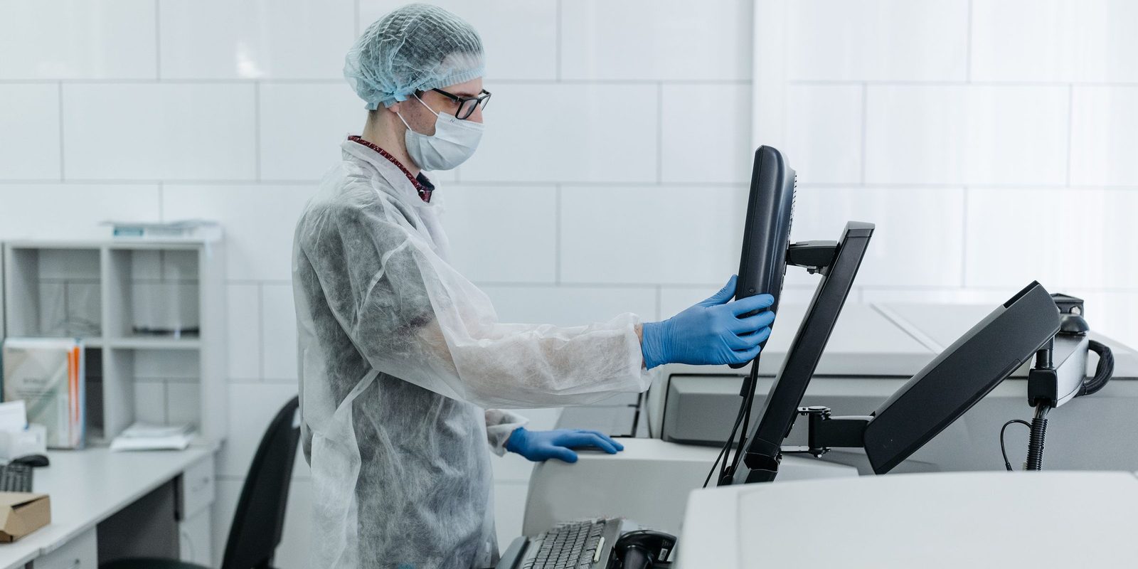 A technician in a disposable coat, hairnet, and mask adjusts a touchscreen terminal in a bright white lab