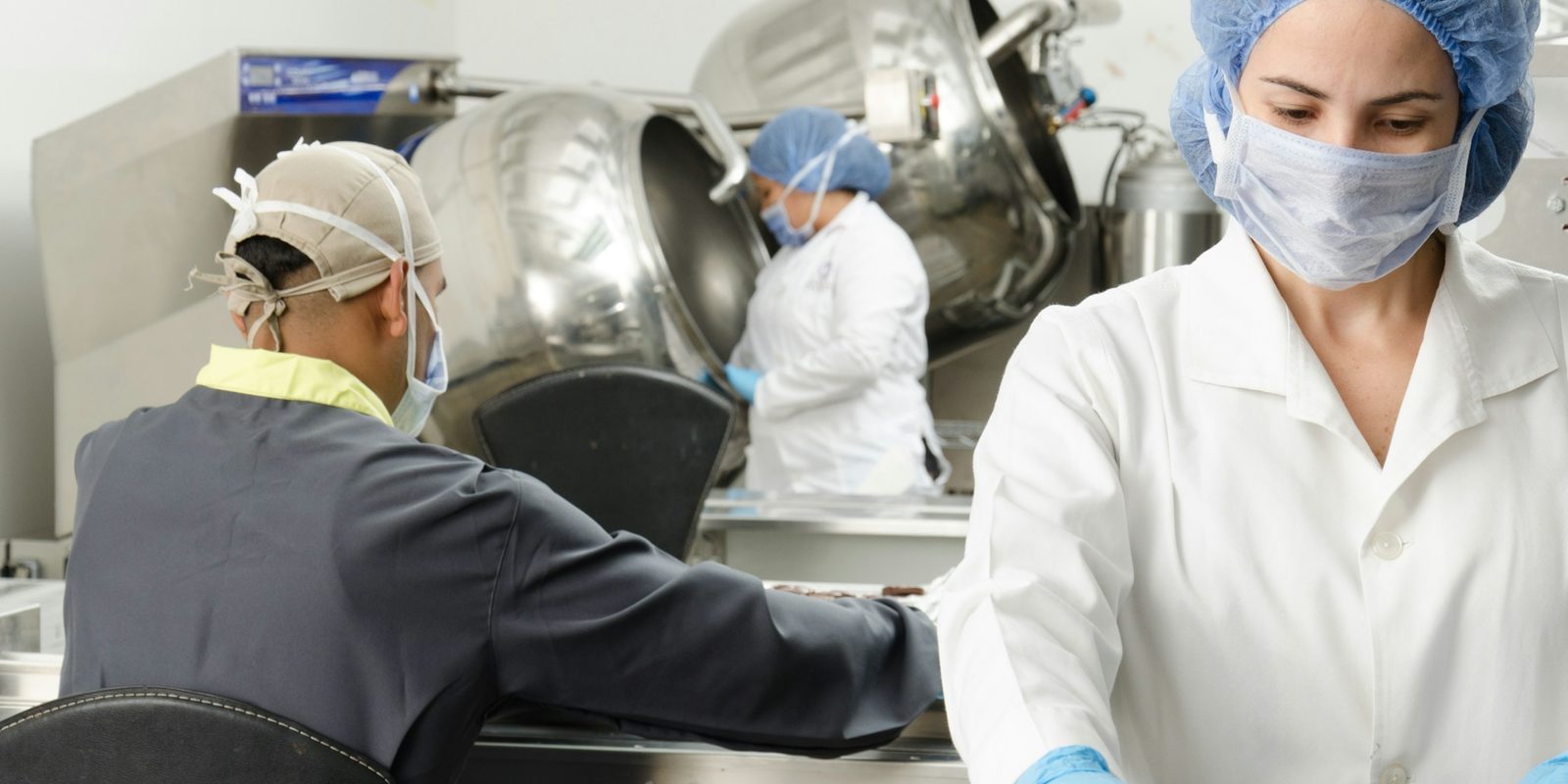 A worker in a hairnet, mask, and white coat works at a conveyor in a food plant, with two colleagues at steel kettles behind her