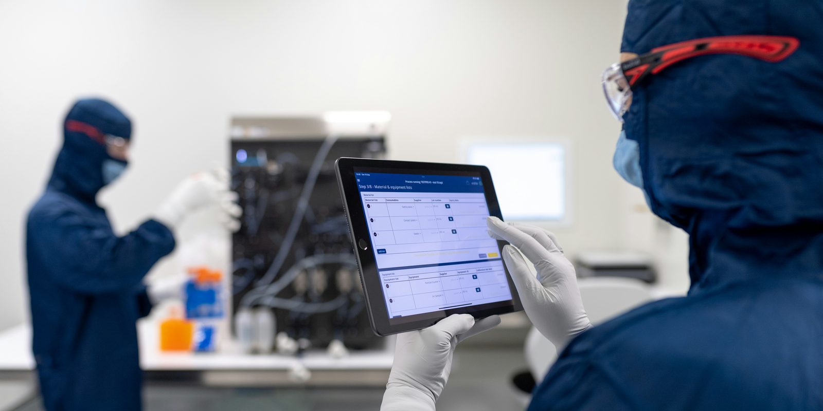 A cleanroom technician in gloves reviews a production checklist on a tablet while a colleague works at equipment