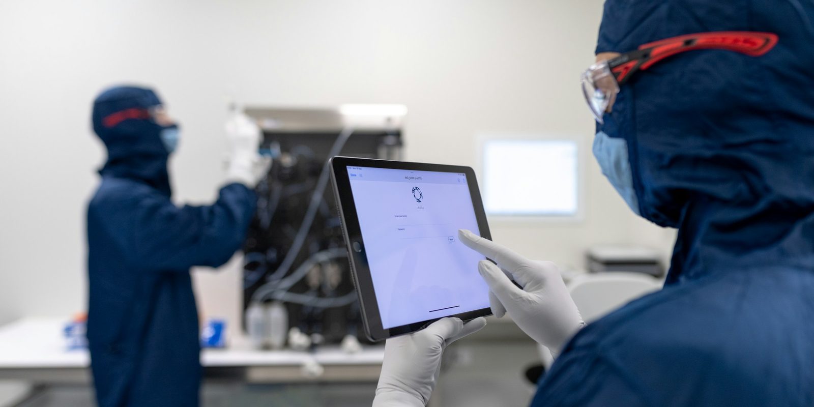 A technician in a blue cleanroom suit and safety glasses reads a tablet while a colleague works at a machine behind