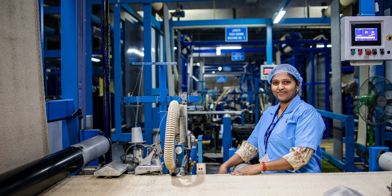 A smiling worker in a blue uniform and hair net stands at a carpet finishing machine, with a control screen on the wall behind