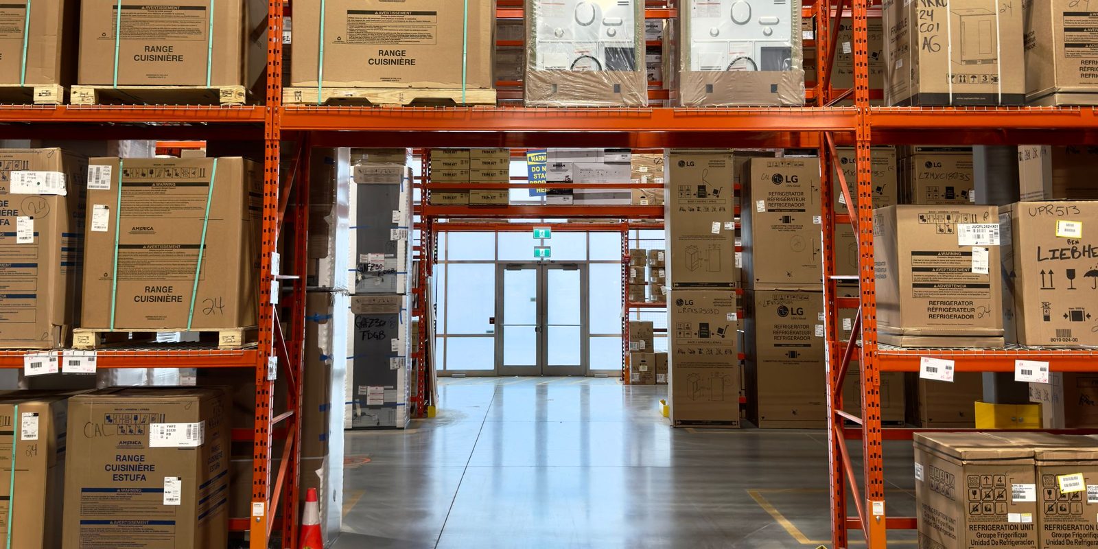 A tidy warehouse with orange pallet racking full of boxed appliances and a clear aisle to the loading doors