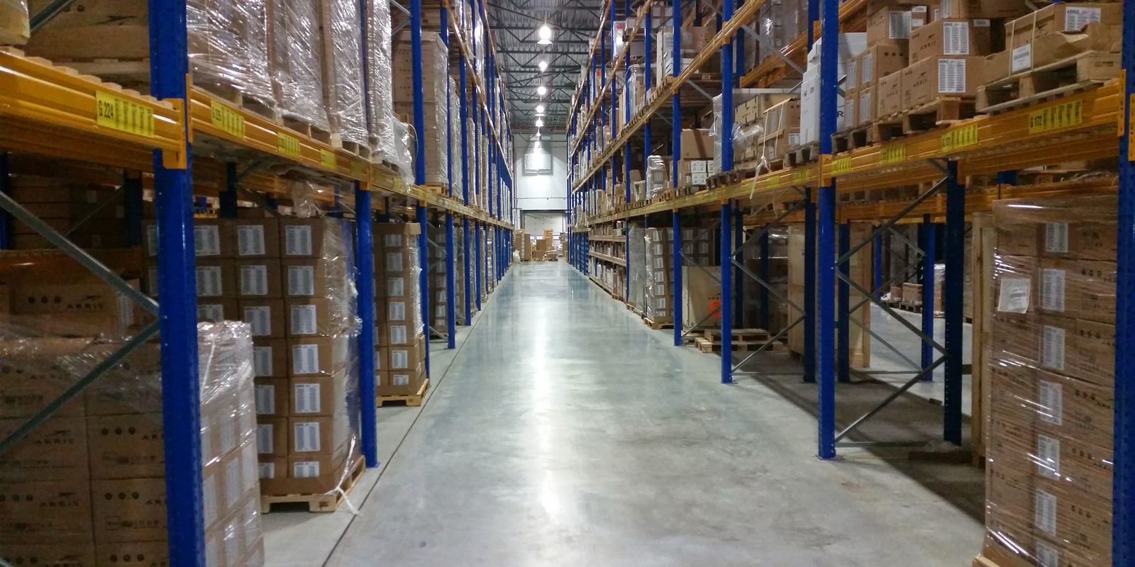 A long, clean aisle between blue and yellow pallet racks loaded with boxed stock in a distribution warehouse