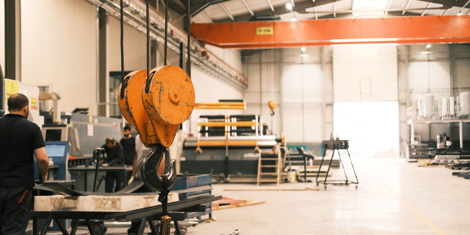 A bright, spacious fabrication hall with an orange overhead crane and hook, workers at machines and a wide clean floor