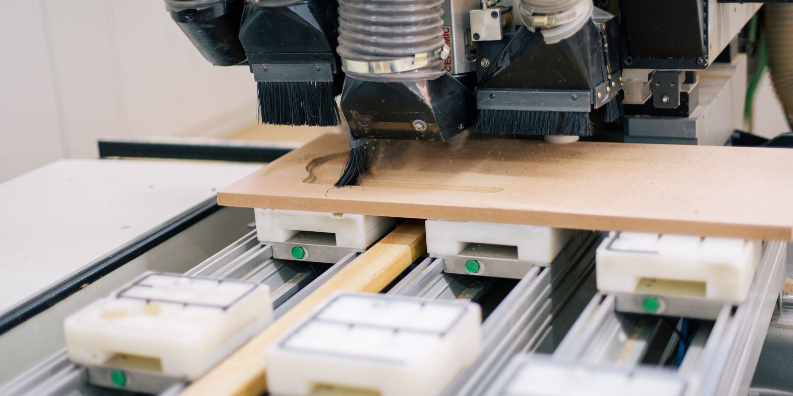 A CNC router head with dust brushes cutting a contour into a wooden panel that sits on white vacuum pods in a bright workshop