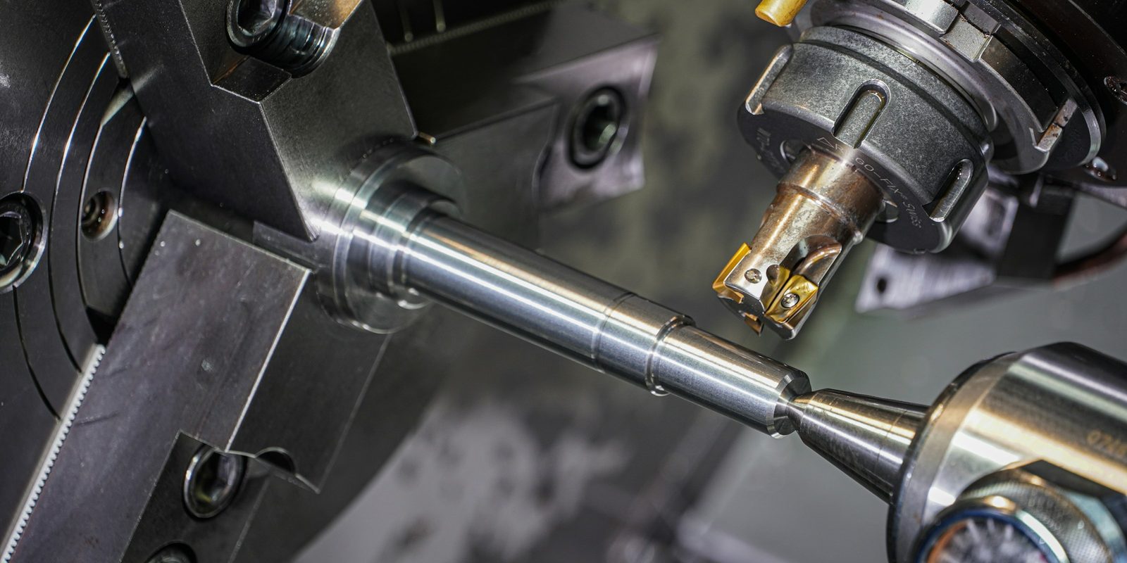 Inside a CNC turning centre, a steel shaft held between the chuck and the tailstock while a milling tool with gold inserts cuts it