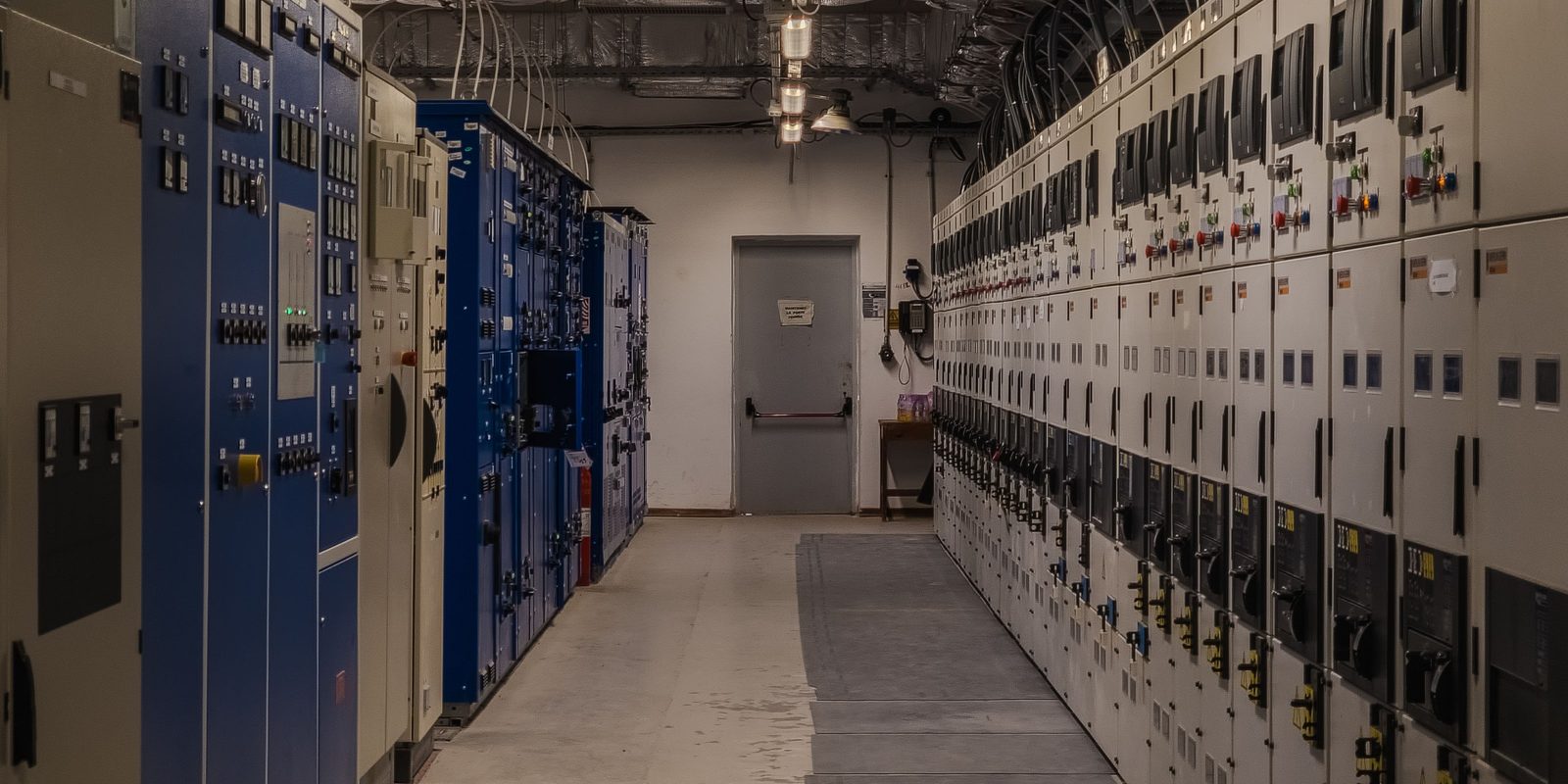 A long, brightly lit switch room lined on both sides with rows of identical grey and blue electrical switchgear cabinets