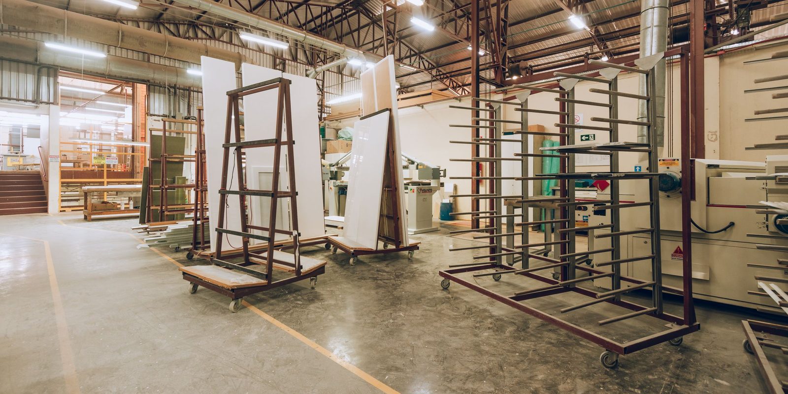 A bright factory workshop with a polished concrete floor, steel trolleys holding white panels, and machines along the back wall