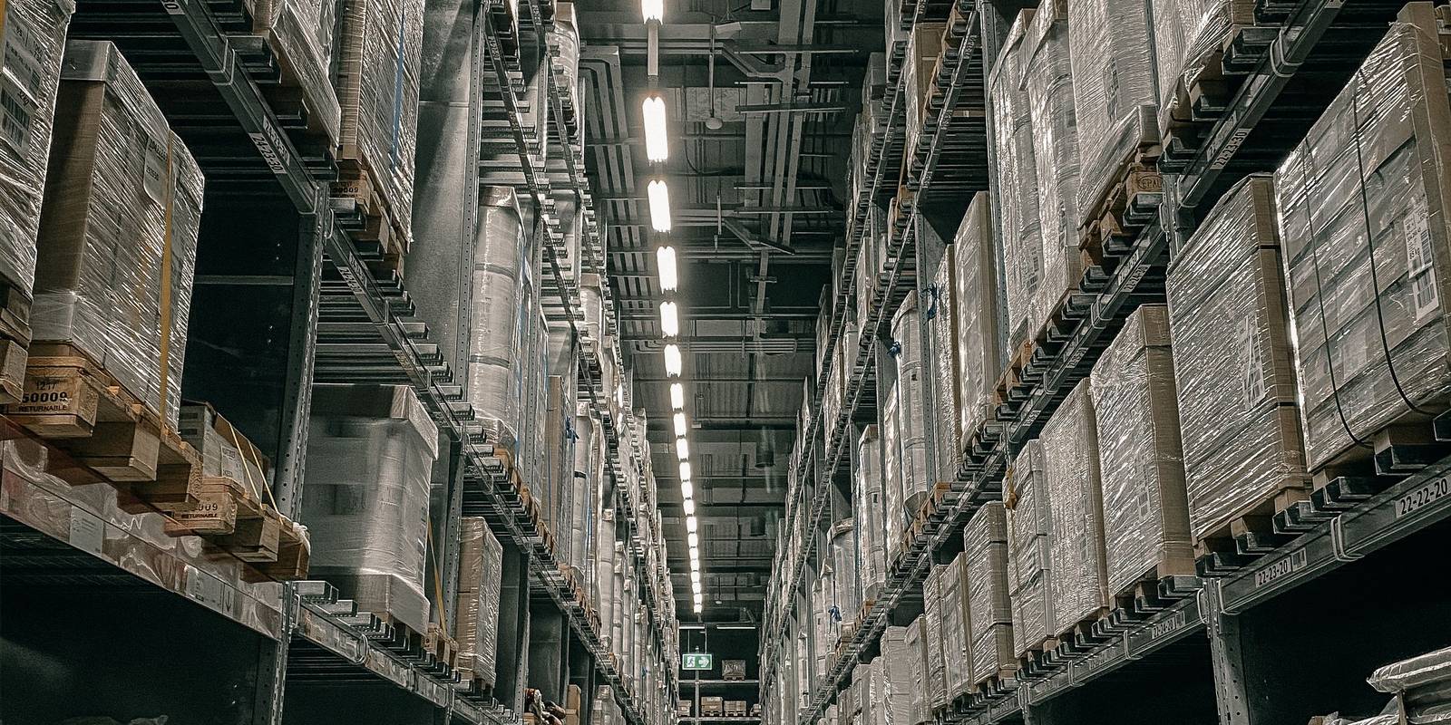 A long warehouse aisle between tall pallet racks loaded with shrink-wrapped cartons, under a line of ceiling lights