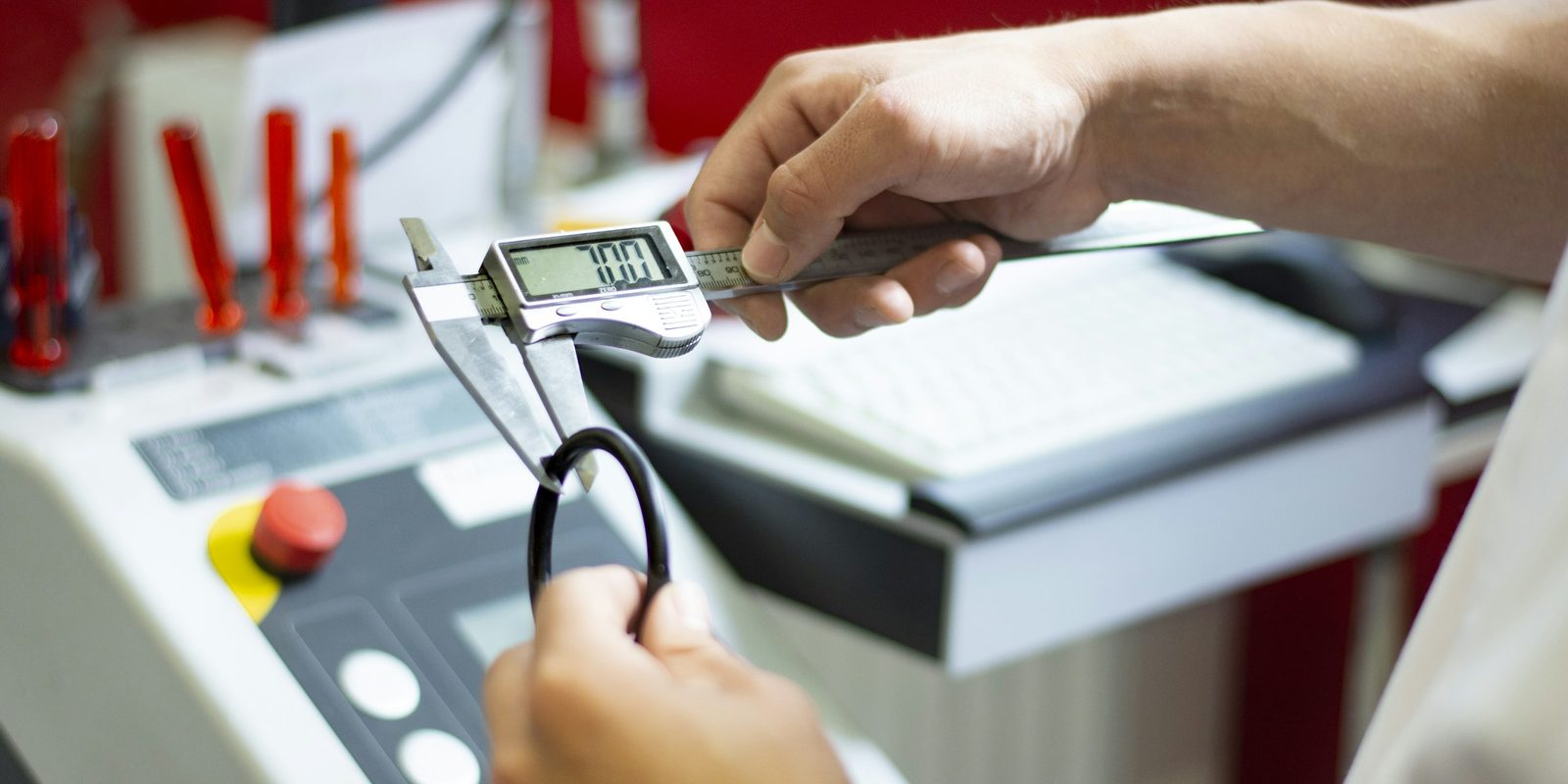 A technician measures a black O-ring with a digital caliper beside the control panel of a white production machine