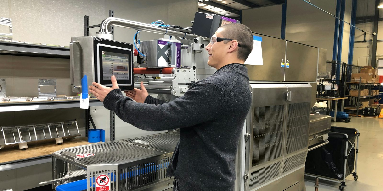 An engineer in safety glasses adjusts the touchscreen control panel of a stainless steel production machine on a bright, tidy plant floor