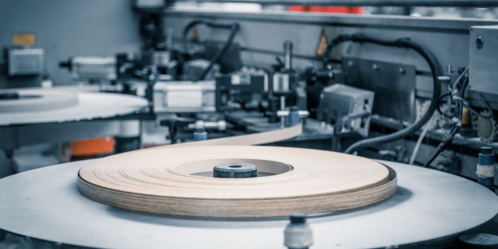 A roll of wood-grain edge banding on the feed table of an edge-banding machine, with guides and cables behind
