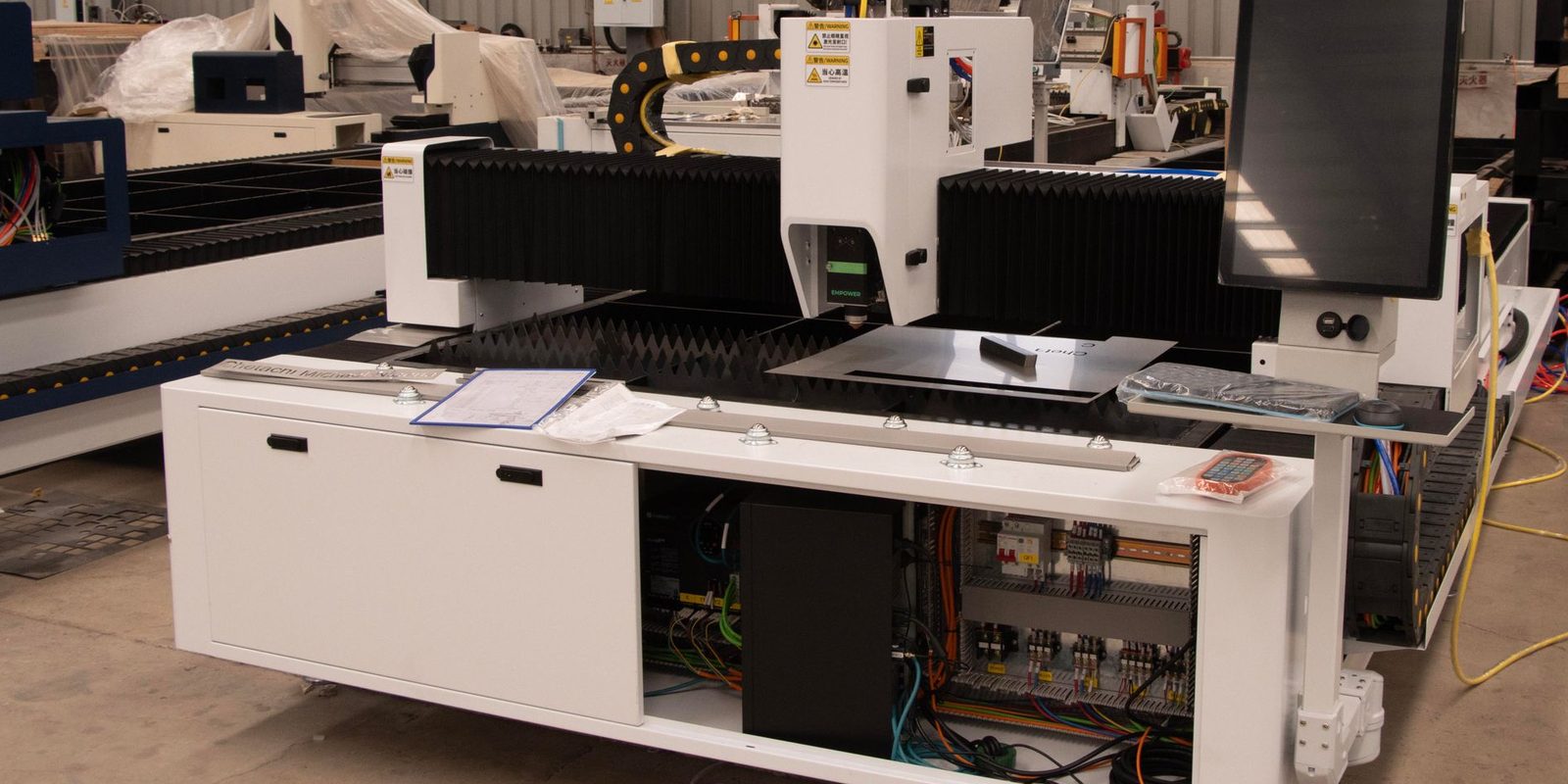 A white laser cutting machine with a large touch screen and an open electrical cabinet on a factory floor