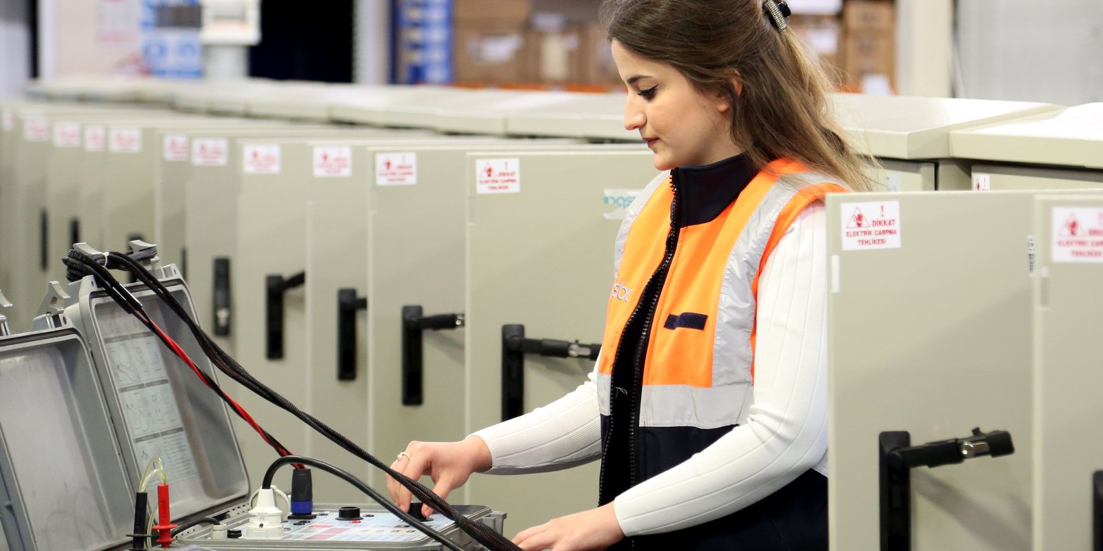 A technician in a high-visibility vest tests a row of new electrical cabinets on a factory floor