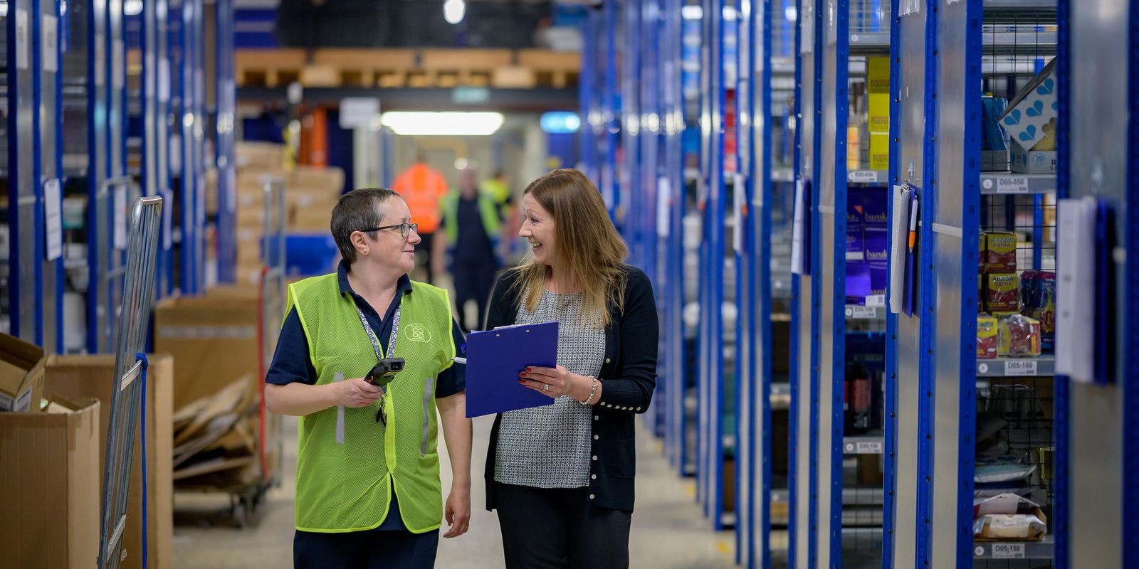 Two warehouse employees walking down an aisle of blue shelving, one holding a handheld scanner and the other a clipboard