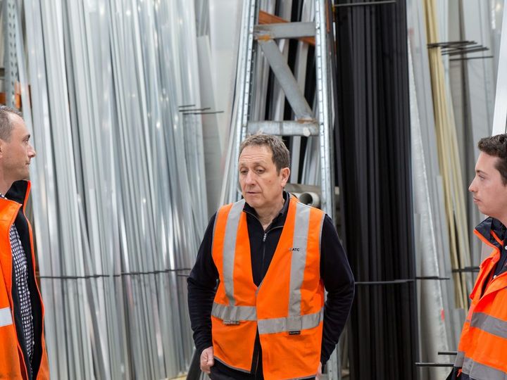 Three people in high-visibility vests talking in a metals warehouse