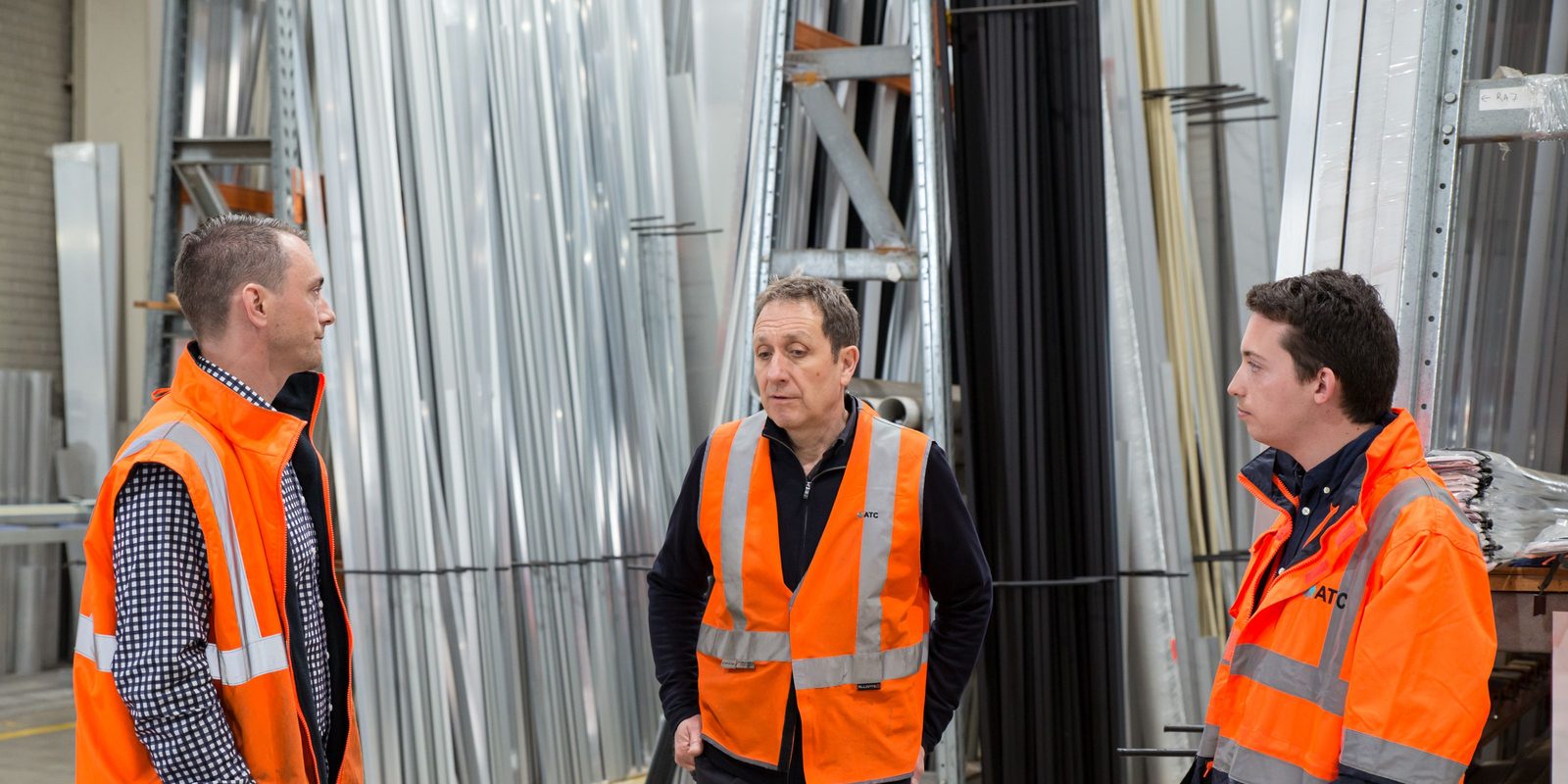 Three men in high-visibility vests talking in a metals warehouse, with racks of aluminum bars behind them