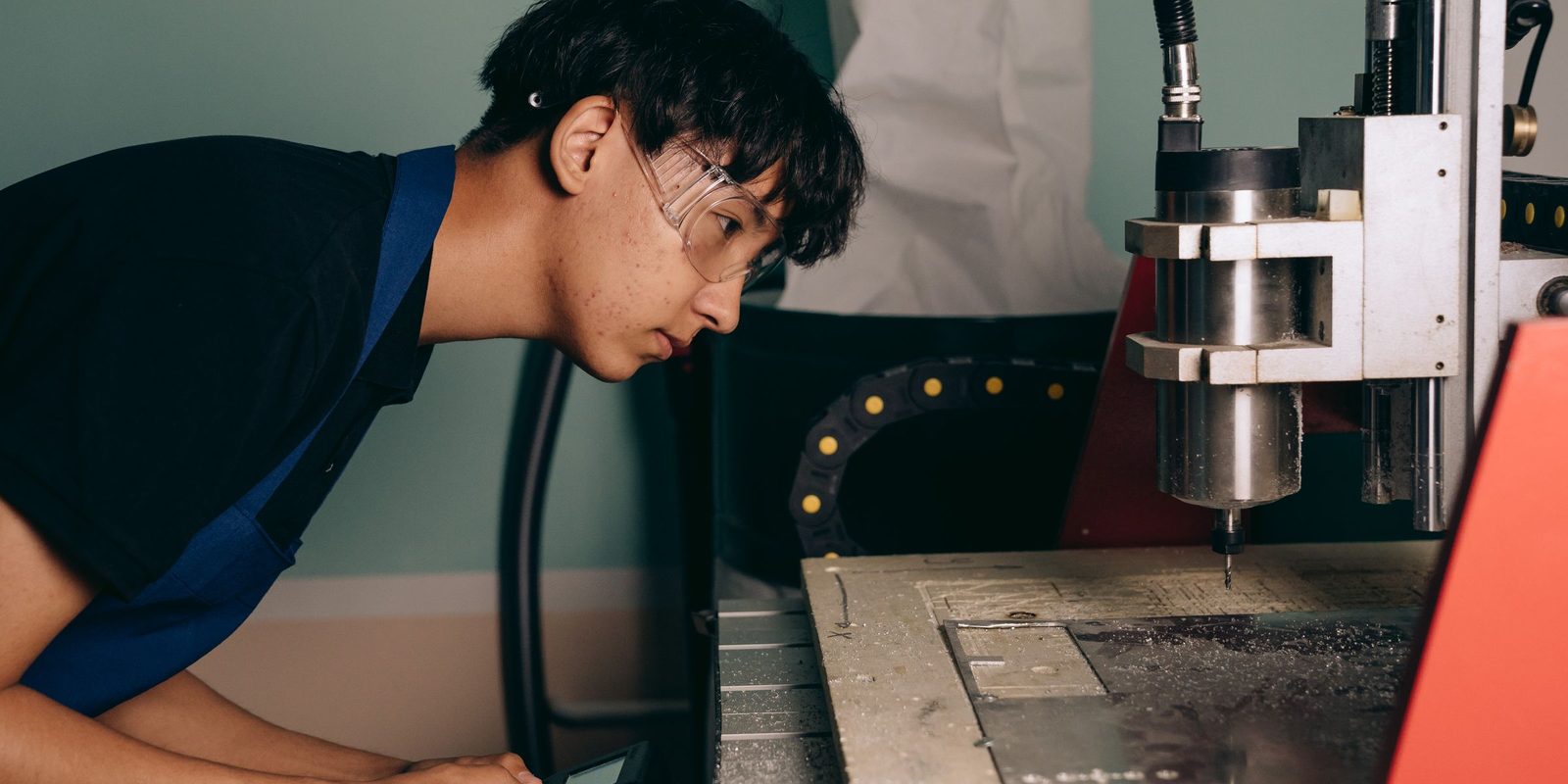 A young machinist in safety glasses and a blue apron watches the spindle of a CNC router cut a metal plate
