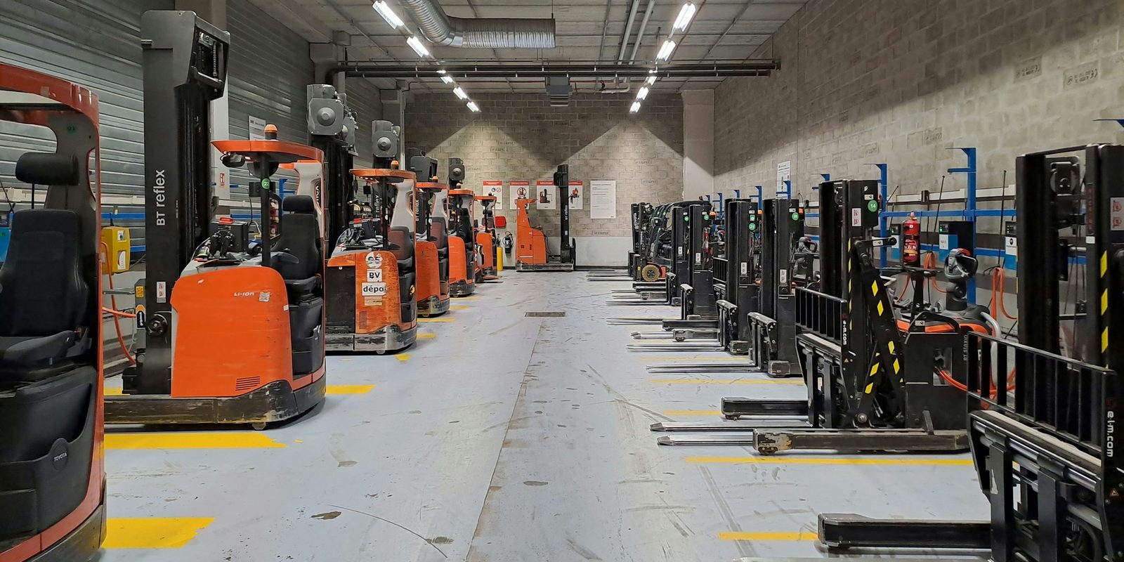 A row of orange reach trucks parked along a bright, clean warehouse aisle under strip lights