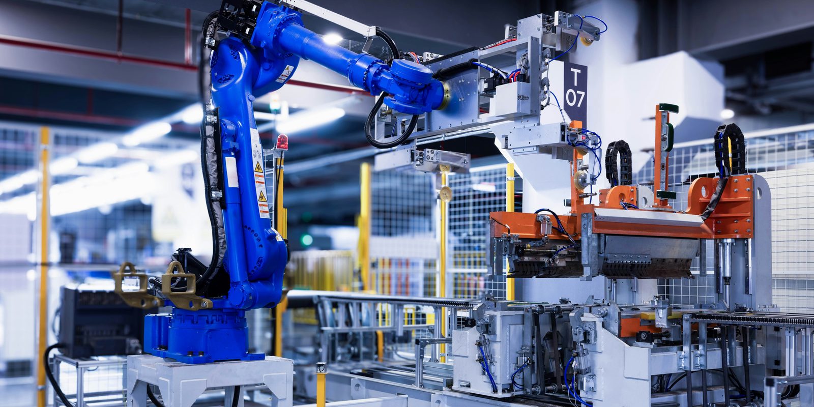 Blue six-axis robot arm beside a fixture in a fenced, brightly lit automated production cell