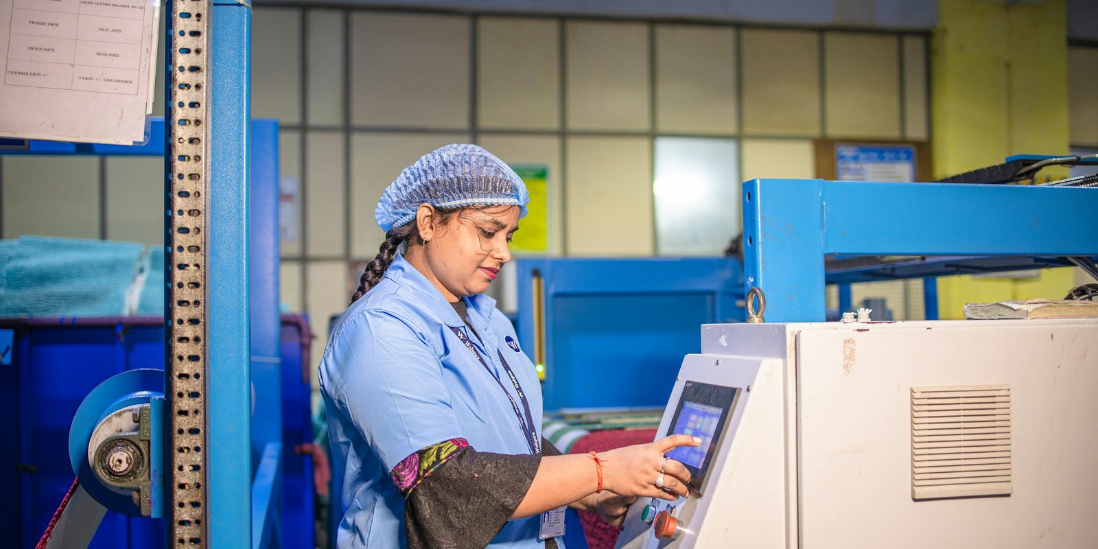 A machine operator in a blue coat and hairnet sets a job on a touchscreen control panel in a bright factory