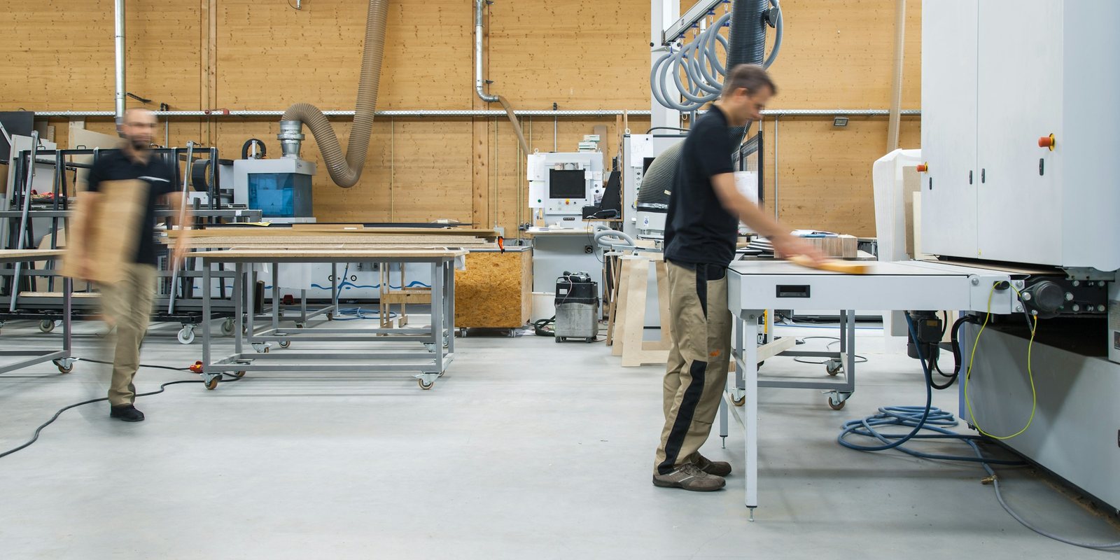 Two workers at long workbenches and a white panel-processing machine in a bright, tidy workshop